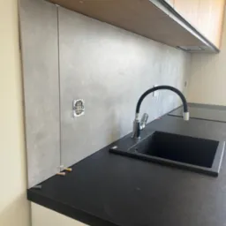 Montaż szarych płytek na ścianie w kuchni, widoczne czarne zlewozmywak i bateria oraz gniazdko elektryczne bez osprzętu, poziomowanie płytek za pomocą klinów.