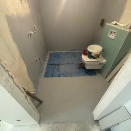 Profesjonalne wykończenia wnętrz Łukasz Szulc - Łazienka w trakcie remontu: wylewka podłogowa, zabezpieczona folią część podłogi, podwieszana toaleta z wiadrem farby na desce, ściany z widocznymi instalacjami i płytą karton gips.