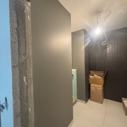 Profesjonalne wykończenia wnętrz Łukasz Szulc - Surowy betonowy oścież w trakcie prac wykończeniowych, ściana pomalowana na szaro, w tle kartony i czarne płytki na ścianie, widoczne przewody elektryczne na suficie.
