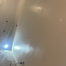 Profesjonalne wykończenia wnętrz Łukasz Szulc - Świeżo pomalowana ściana w jasnym odcieniu, widoczny wałek malarski w wiadrze z farbą, przewody elektryczne wystające ze ściany, drabina oparta o ścianę i lampa oświetlająca miejsce pracy.