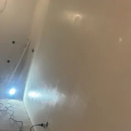 Świeżo pomalowana ściana w jasnym odcieniu, widoczny wałek malarski w wiadrze z farbą, przewody elektryczne wystające ze ściany, drabina oparta o ścianę i lampa oświetlająca miejsce pracy.