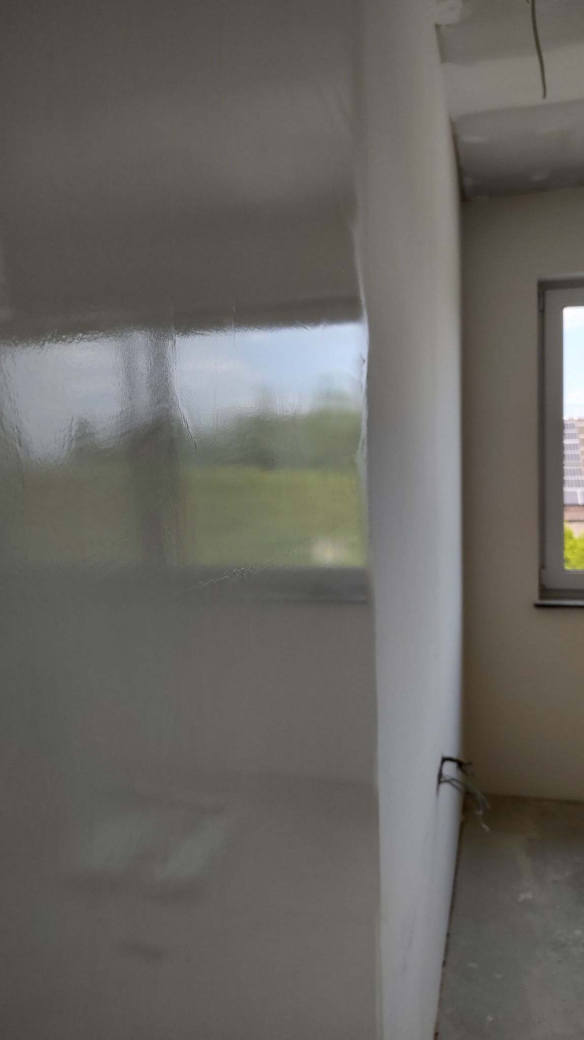 Świeżo nałożona, gładka, szara powłoka na ścianie w nowym pomieszczeniu, z widocznym odbiciem okna i krajobrazu za nim, oraz wystającymi kablami elektrycznymi przy podłodze.