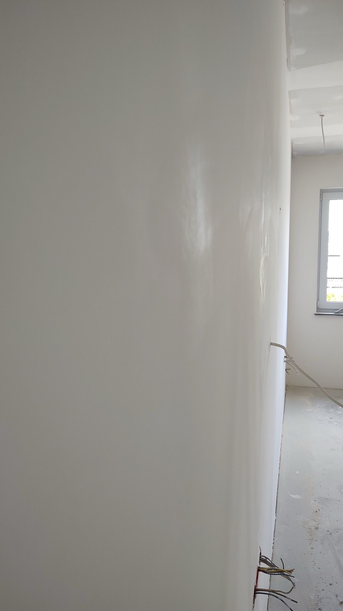 Świeżo położona, jasna, prawie gładka ściana we Wrocławiu, z widocznymi przewodami elektrycznymi wystającymi ze ściany przy podłodze, w tle okno i surowy stan wykończenia.