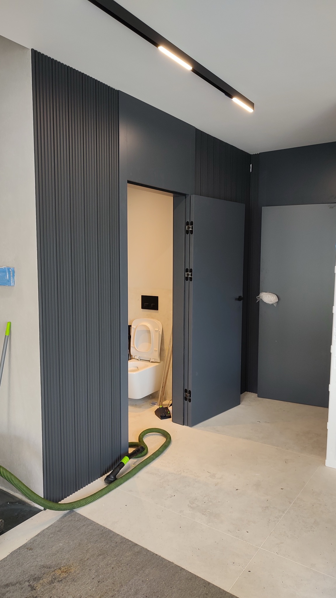 Wnętrze łazienki w trakcie wykańczania z widoczną toaletą, szarą boazerią ścienną, drzwiami i rurą odkurzacza przemysłowego na podłodze.