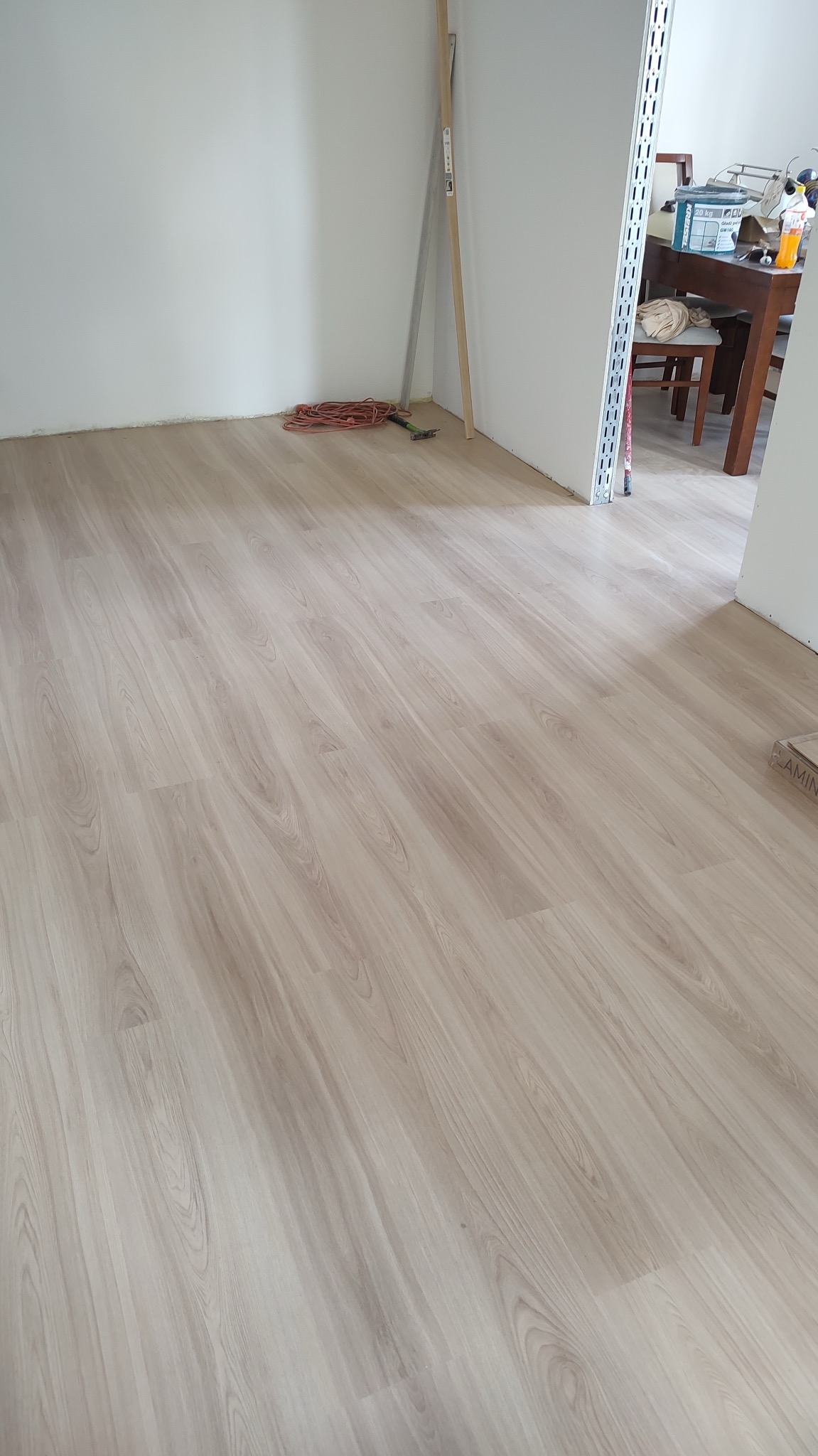 Nowo położona podłoga laminowana o jasnym, drewnopodobnym wzorze w pustym pokoju z białymi ścianami; widoczne narzędzia i zwinięty przedłużacz w rogu, sugerujące trwające prace wykończeniowe.