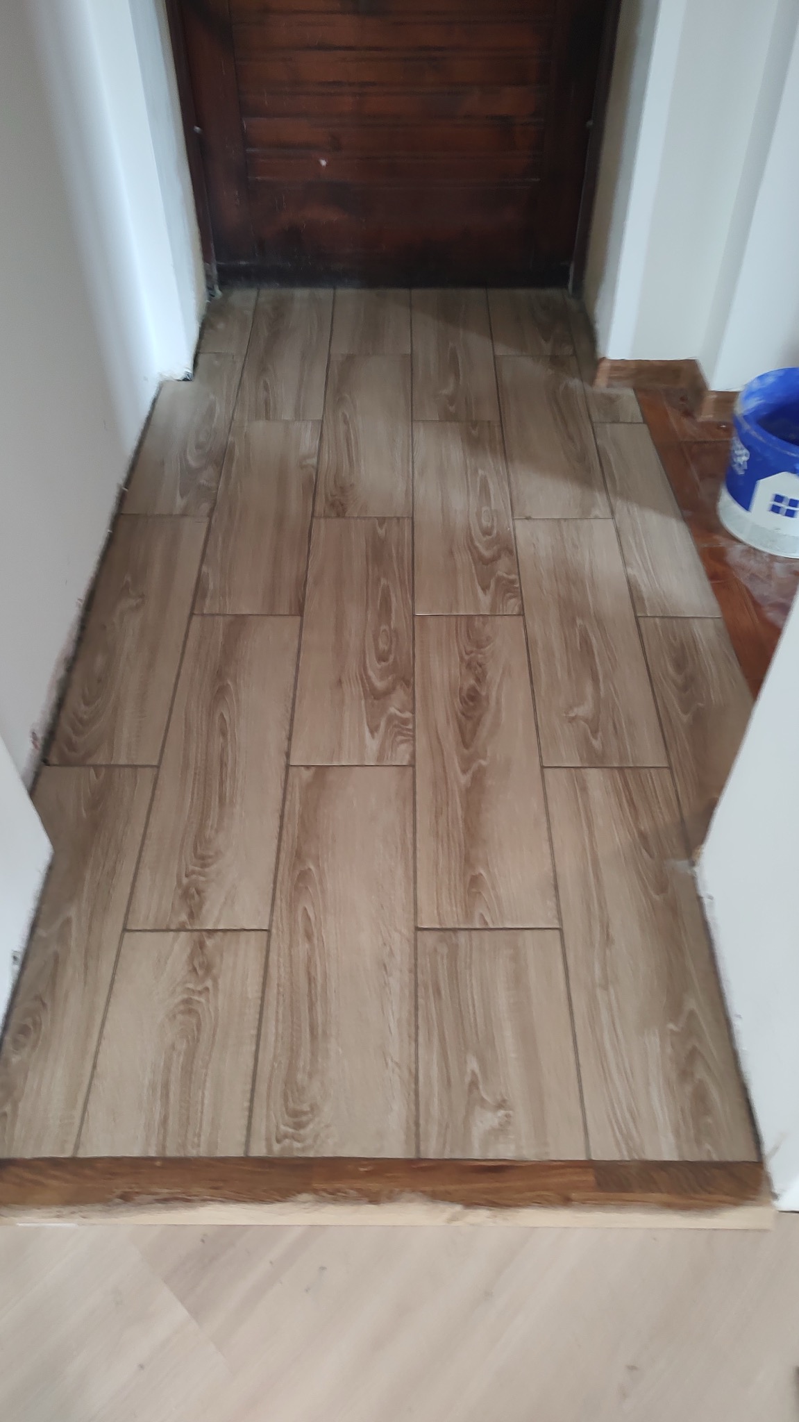 Ułożona podłoga z płytek imitujących drewno w przejściu między pomieszczeniami, widoczna listwa progowa i fragment wiadra z farbą.