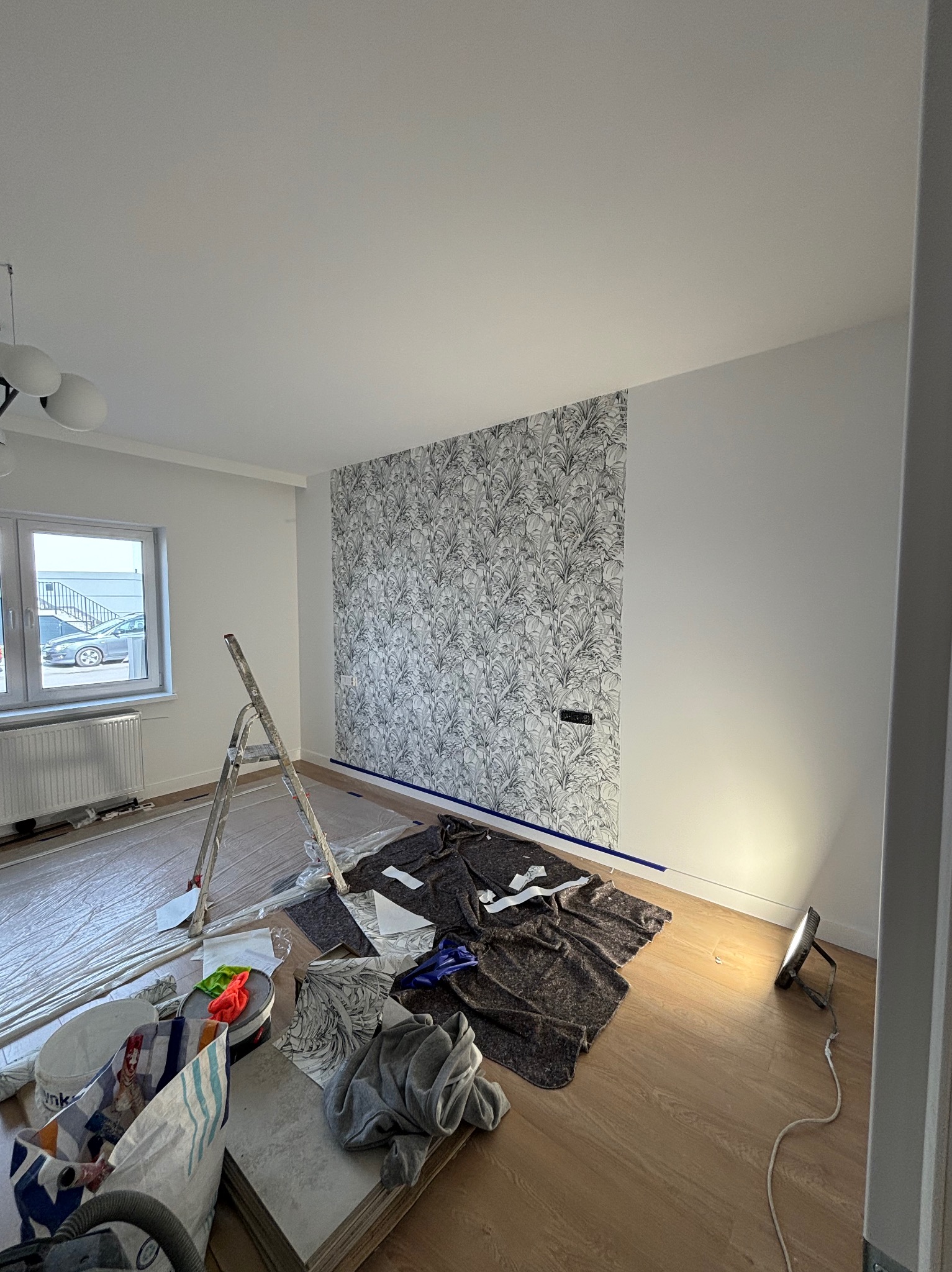 Pokój w trakcie remontu: częściowo wytapetowana ściana w roślinny wzór, zabezpieczona podłoga, drabina oparta o ścianę, lampa halogenowa oświetlająca fragment podłogi, widoczne narzędzia malarskie...