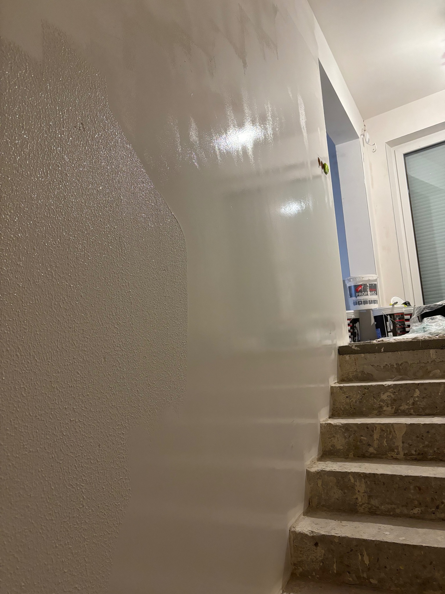 Ściana po częściowym nałożeniu gładzi, widoczna różnica w strukturze powierzchni, w tle puszki z gładzią na schodach.