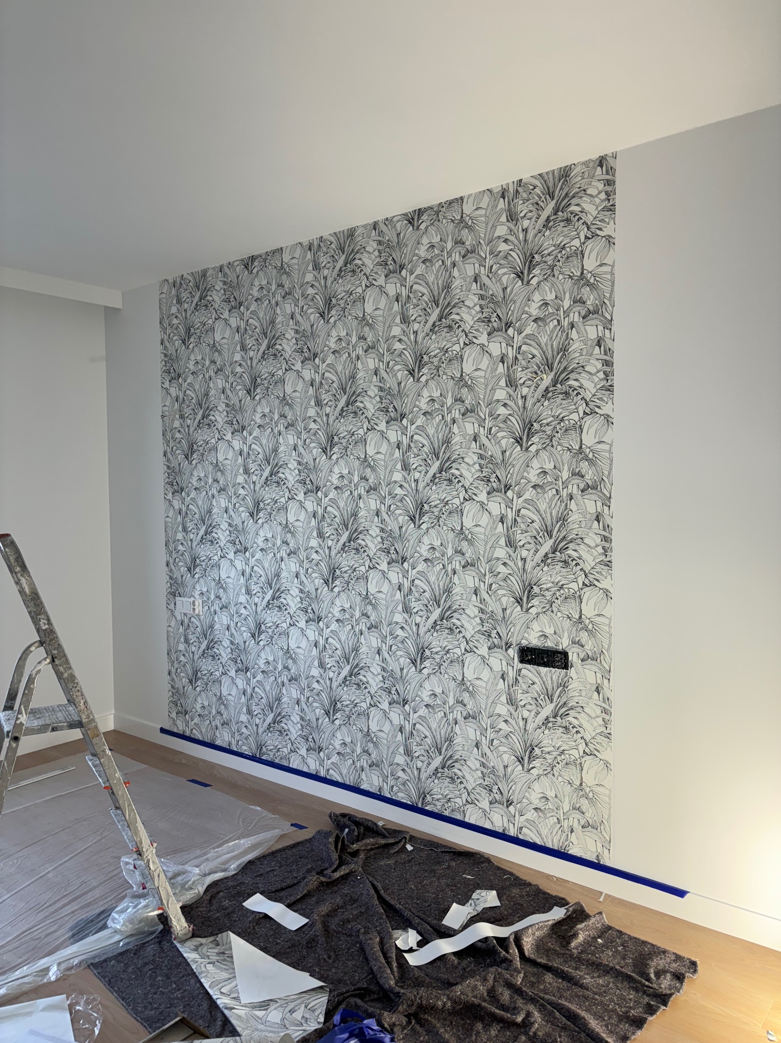 Fragment ściany z nowo położoną tapetą w tropikalny wzór, zabezpieczona niebieską taśmą malarską, z widoczną drabiną i folią ochronną na podłodze podczas prac.