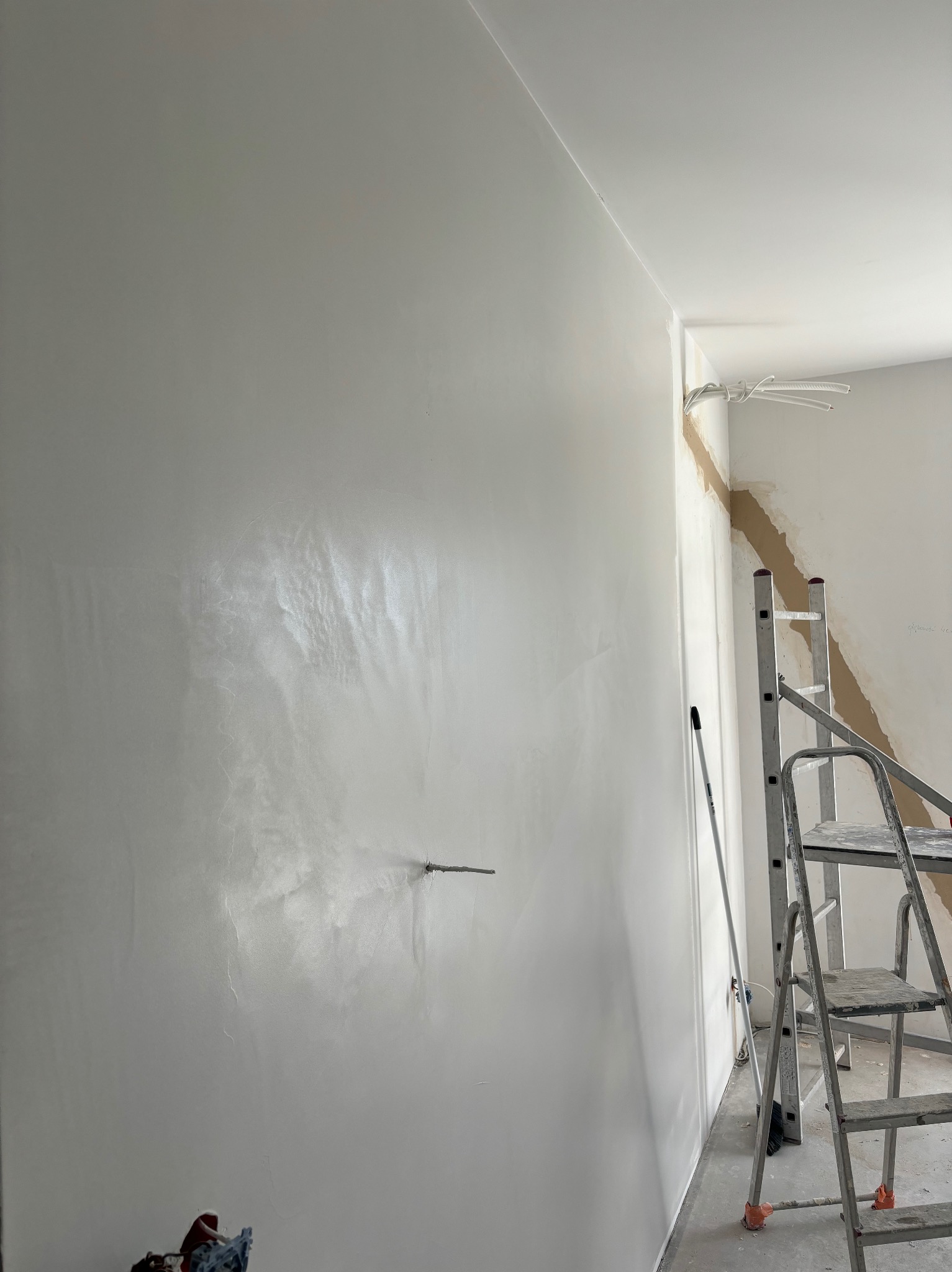 Ściana po nałożeniu gładzi szpachlowej, widoczne delikatne refleksy światła, w rogu drabina malarska i wystające kable elektryczne.