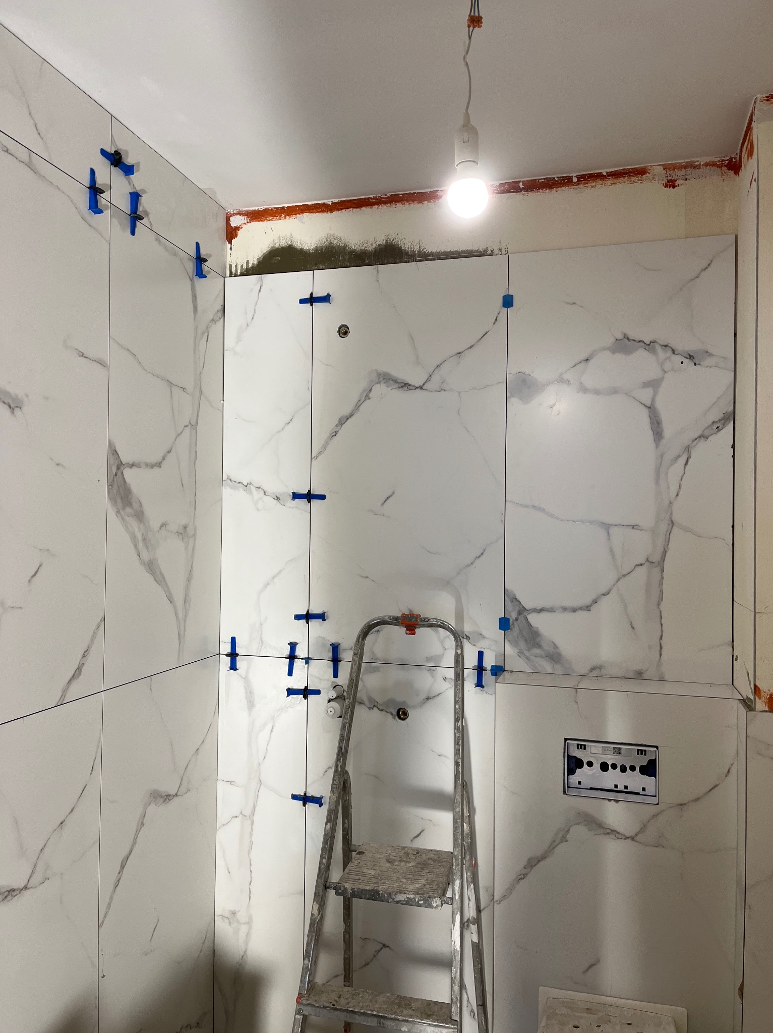 Ściana łazienki w trakcie układania białych płytek imitujących marmur, z użyciem niebieskich klipsów poziomujących, widoczna drabina malarska i wystająca żarówka.