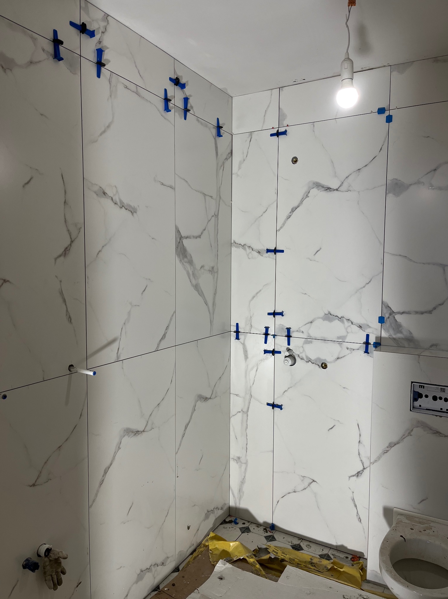 Świeżo ułożone, białe płytki imitujące marmur na ścianach łazienki, z użyciem niebieskich klipsów poziomujących, widoczny fragment instalacji sanitarnej i surowy stan wykończenia.