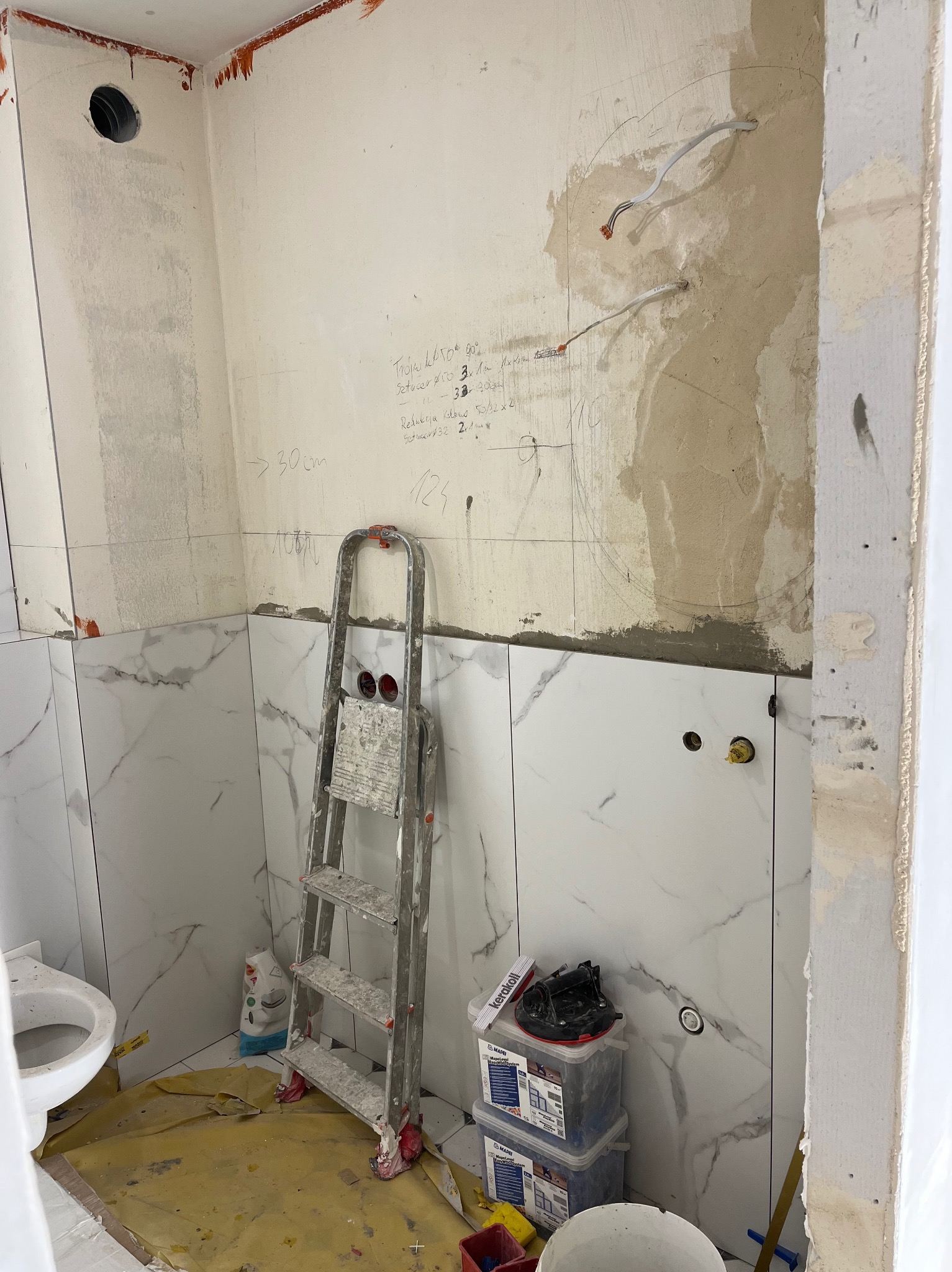 Remont łazienki w trakcie: ułożone marmurowe płytki częściowo zakrywają ściany z instalacją elektryczną i notatkami, drabina monterska oparta o ścianę, widoczne narzędzia i materiały budowlane.