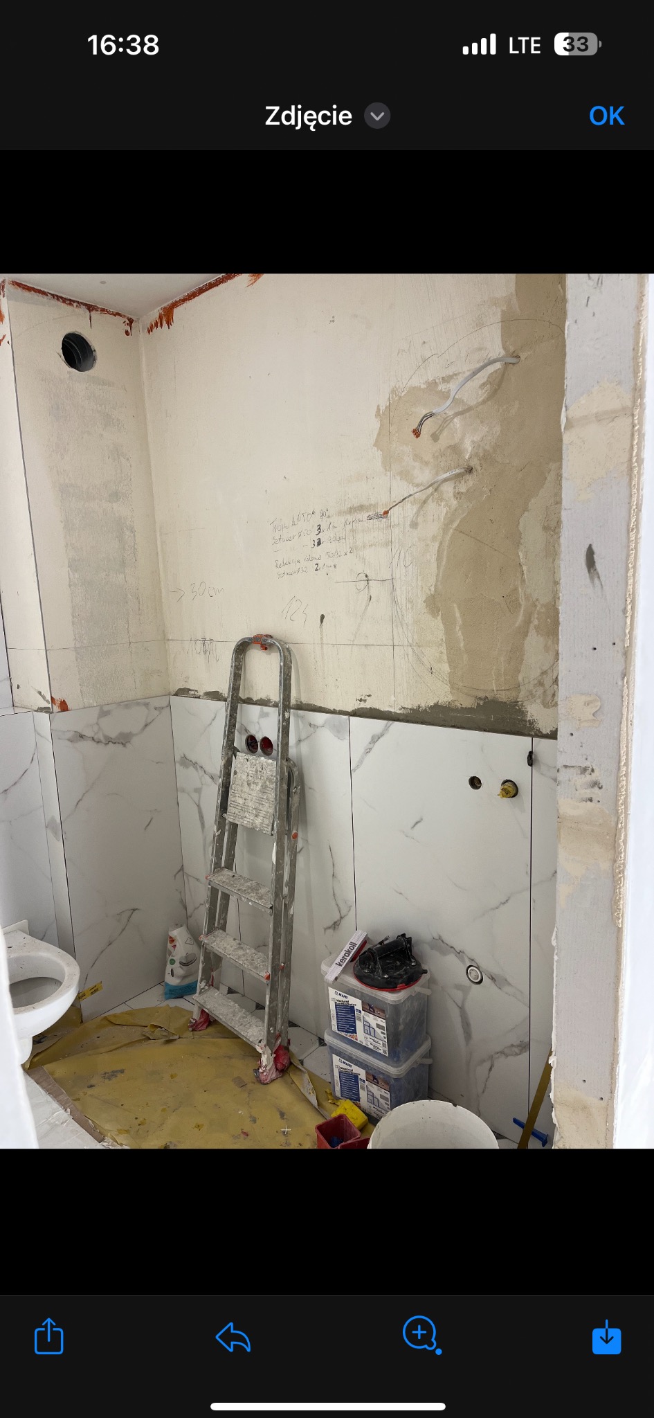 Łazienka w trakcie remontu: częściowo ułożone białe płytki imitujące marmur na ścianach, widoczne ślady po instalacjach elektrycznych, drabina malarska, pojemniki z zaprawą, przygotowane podłoże...