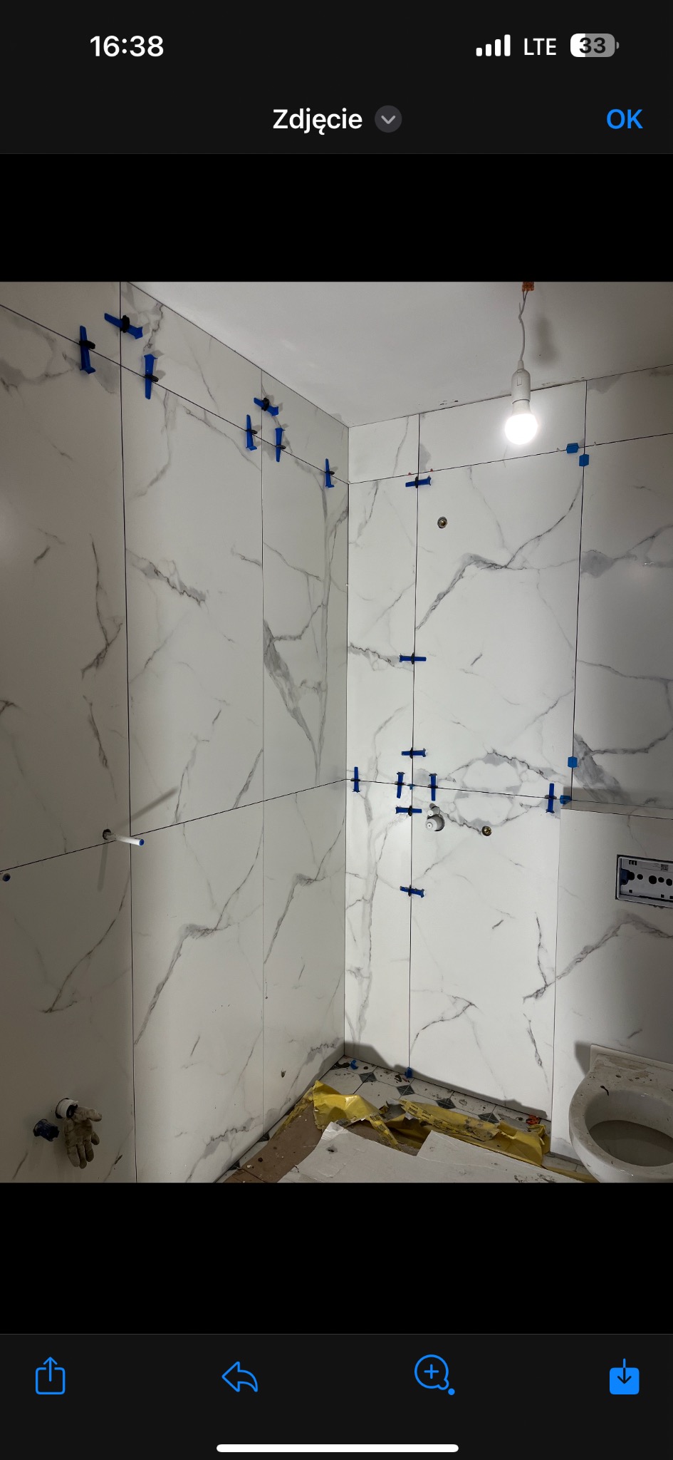 Białe płytki imitujące marmur na ścianach łazienki, z niebieskimi klipsami poziomującymi i czarną linką wyznaczającą linie, widoczna instalacja pod umywalkę i fragment toalety w trakcie remontu.