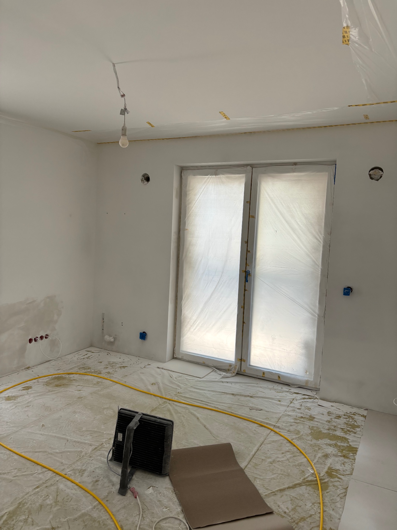 Pokój w trakcie prac przygotowawczych do malowania: okna zabezpieczone folią, podłoga przykryta folią ochronną, widoczne puszki elektryczne w ścianach, lampa wisząca na kablu, grzejnik elektryczny...