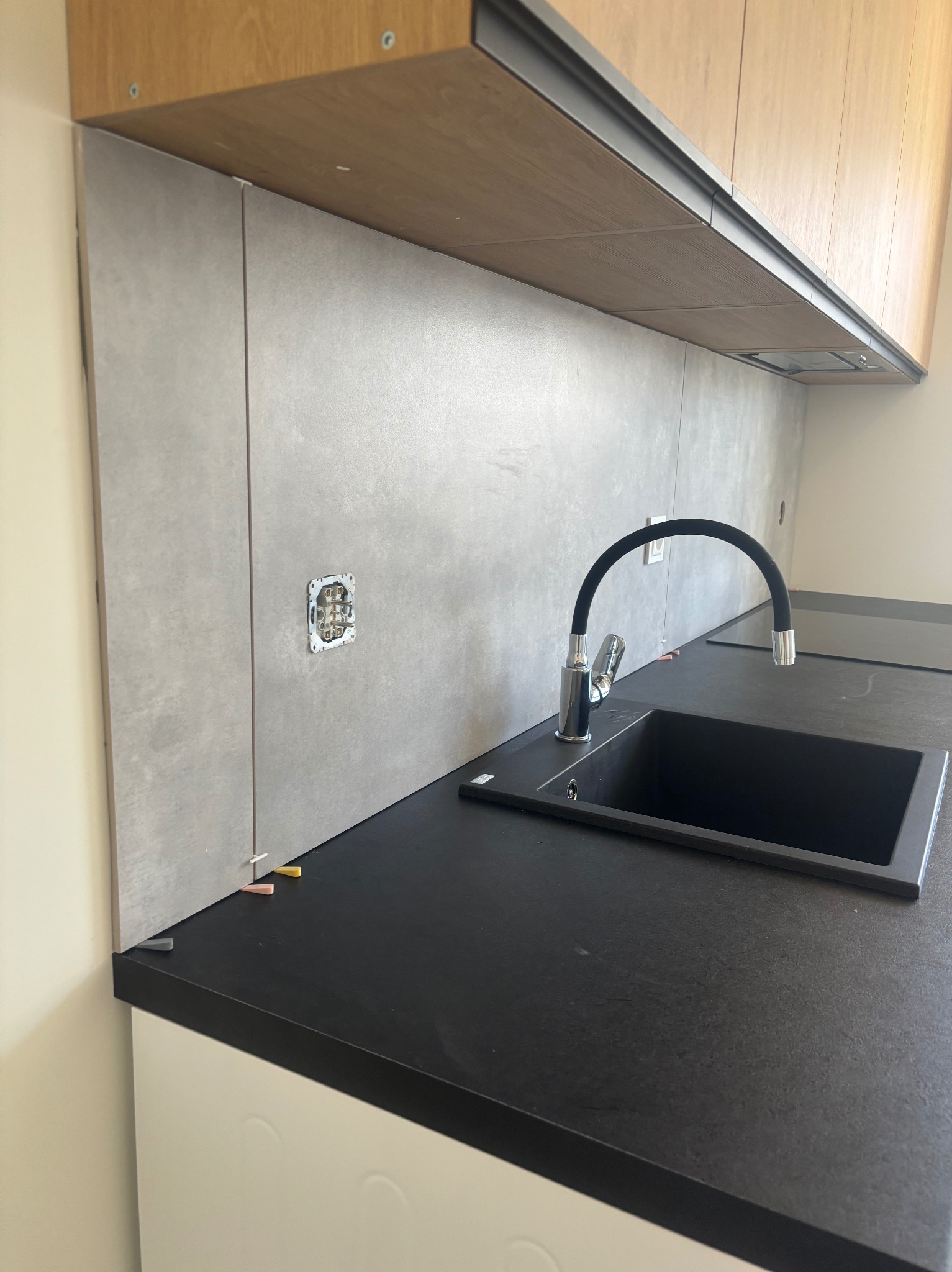 Montaż szarych płytek na ścianie w kuchni, widoczne czarne zlewozmywak i bateria oraz gniazdko elektryczne bez osprzętu, poziomowanie płytek za pomocą klinów.