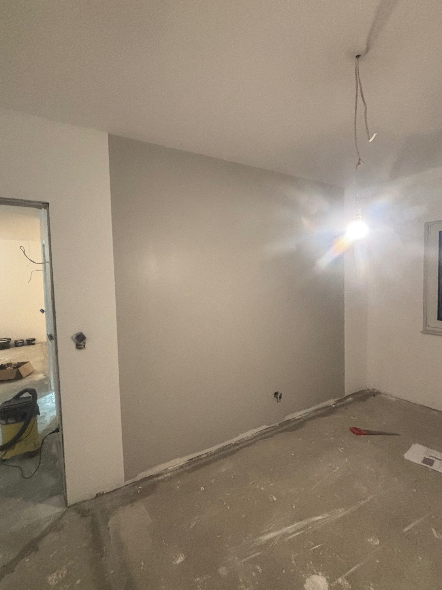 Pokój w trakcie prac przygotowawczych: ściany częściowo pomalowane na szaro, surowa podłoga, widoczne przewody elektryczne i narzędzia malarskie.