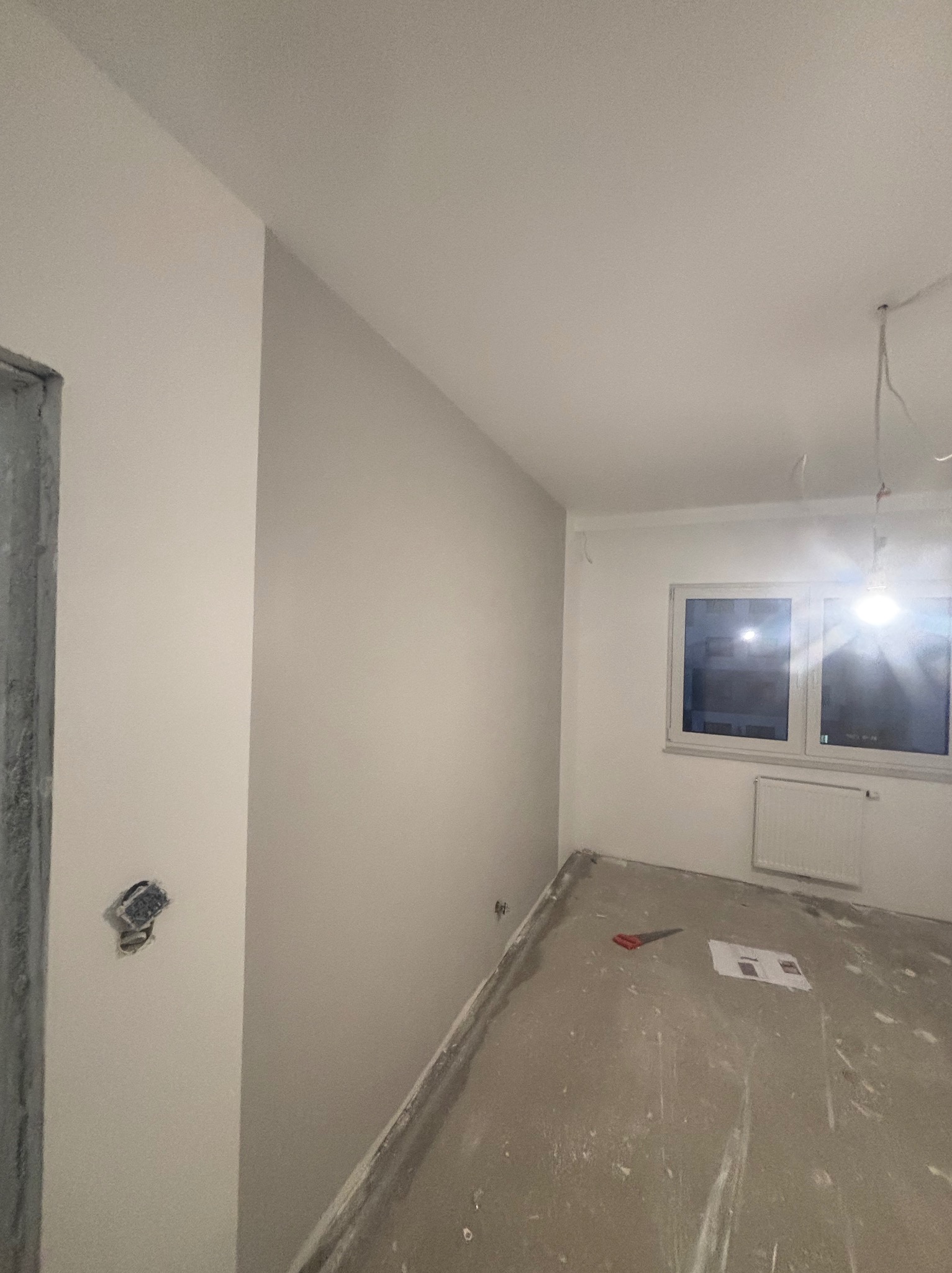 Świeżo pomalowane, jasne wnętrze z widocznym gniazdkiem elektrycznym bez osprzętu, przygotowane do dalszych prac wykończeniowych, z narzędziami i planami leżącymi na surowej podłodze.