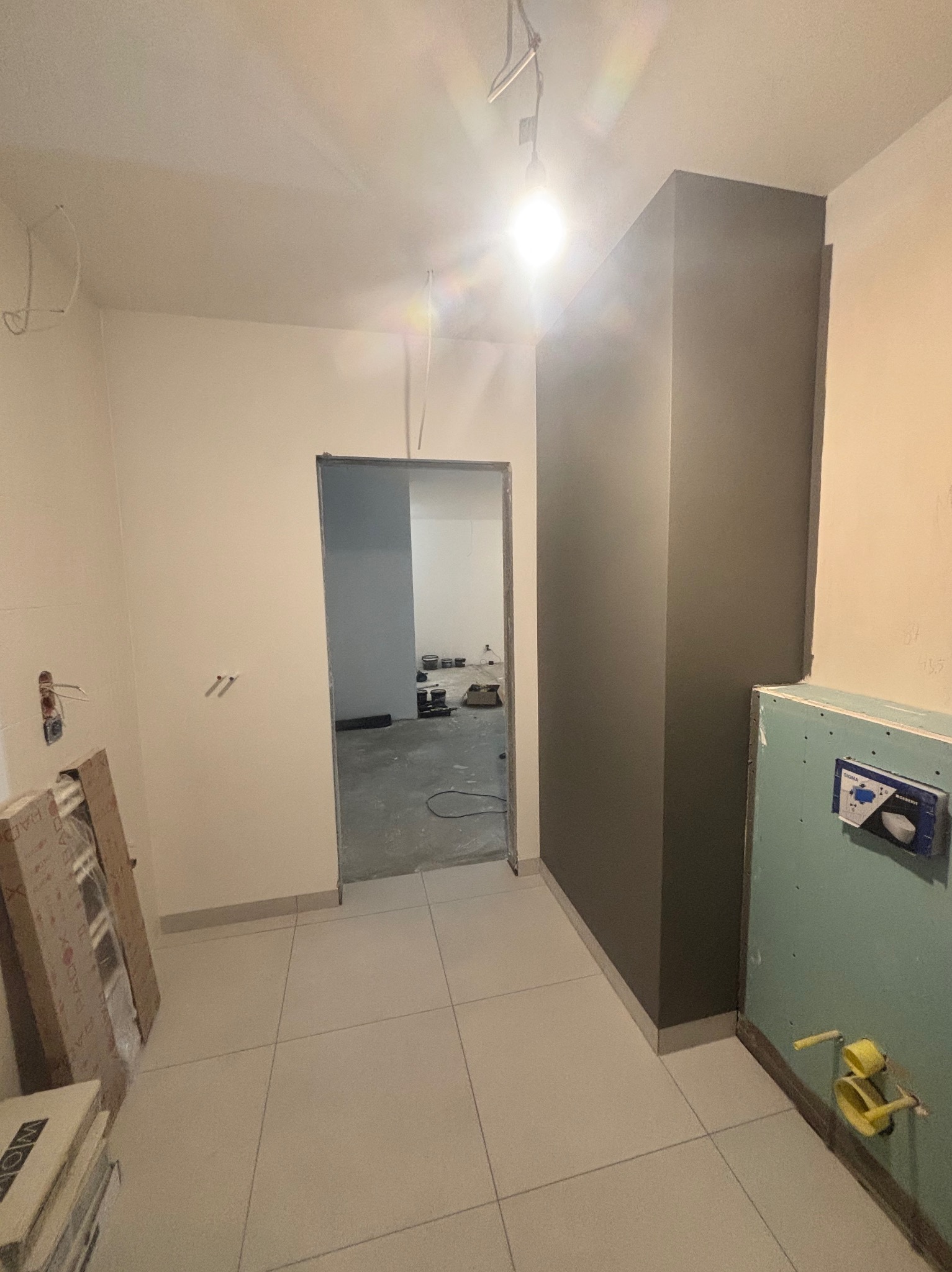 Wnętrze w trakcie prac wykończeniowych: widoczna szara kolumna, częściowo obudowany stelaż WC z płyt karton gips, surowe ściany z wyprowadzonymi instalacjami, skrzynki z płytkami pod ścianą...