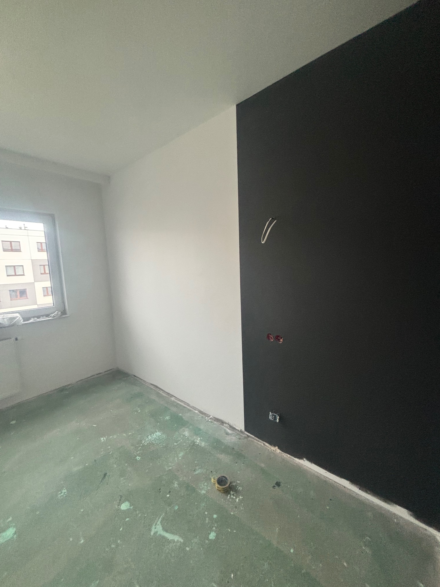 Pokój po częściowym pomalowaniu ścian na biało i czarno, widoczne puszki elektryczne i wystające przewody, podłoga zabezpieczona folią malarską.