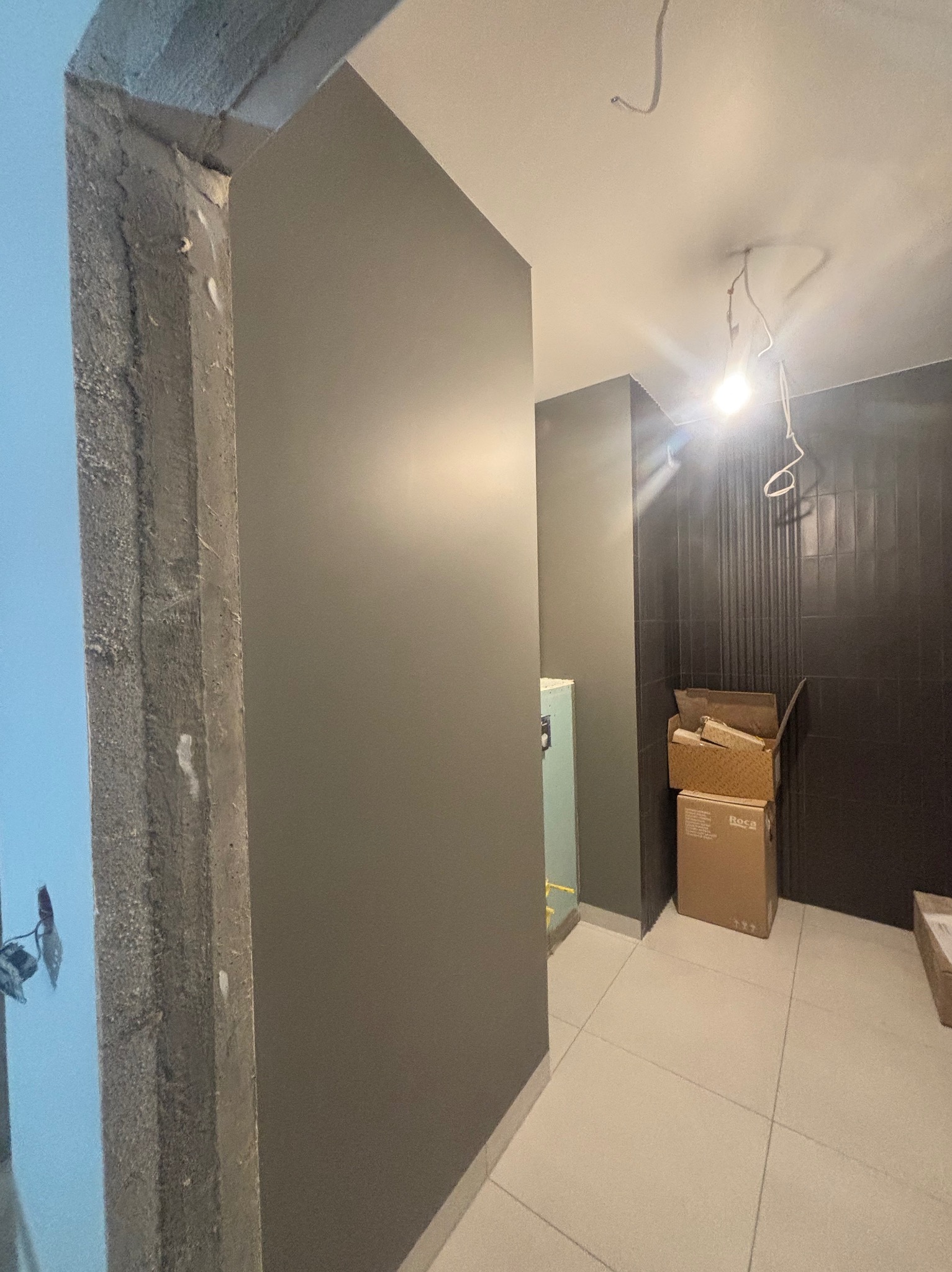 Surowy betonowy oścież w trakcie prac wykończeniowych, ściana pomalowana na szaro, w tle kartony i czarne płytki na ścianie, widoczne przewody elektryczne na suficie.