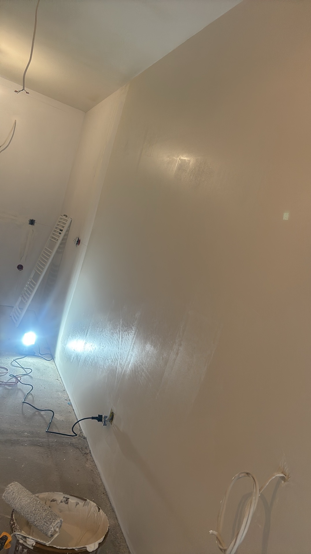 Świeżo pomalowana ściana w jasnym odcieniu, widoczny wałek malarski w wiadrze z farbą, przewody elektryczne wystające ze ściany, drabina oparta o ścianę i lampa oświetlająca miejsce pracy.