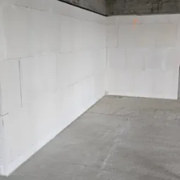 Wnętrze surowego domu w trakcie budowy z białymi ścianami z betonu komórkowego i surowym betonowym stropem, widoczny fragment pomarańczowej linii na ścianie.