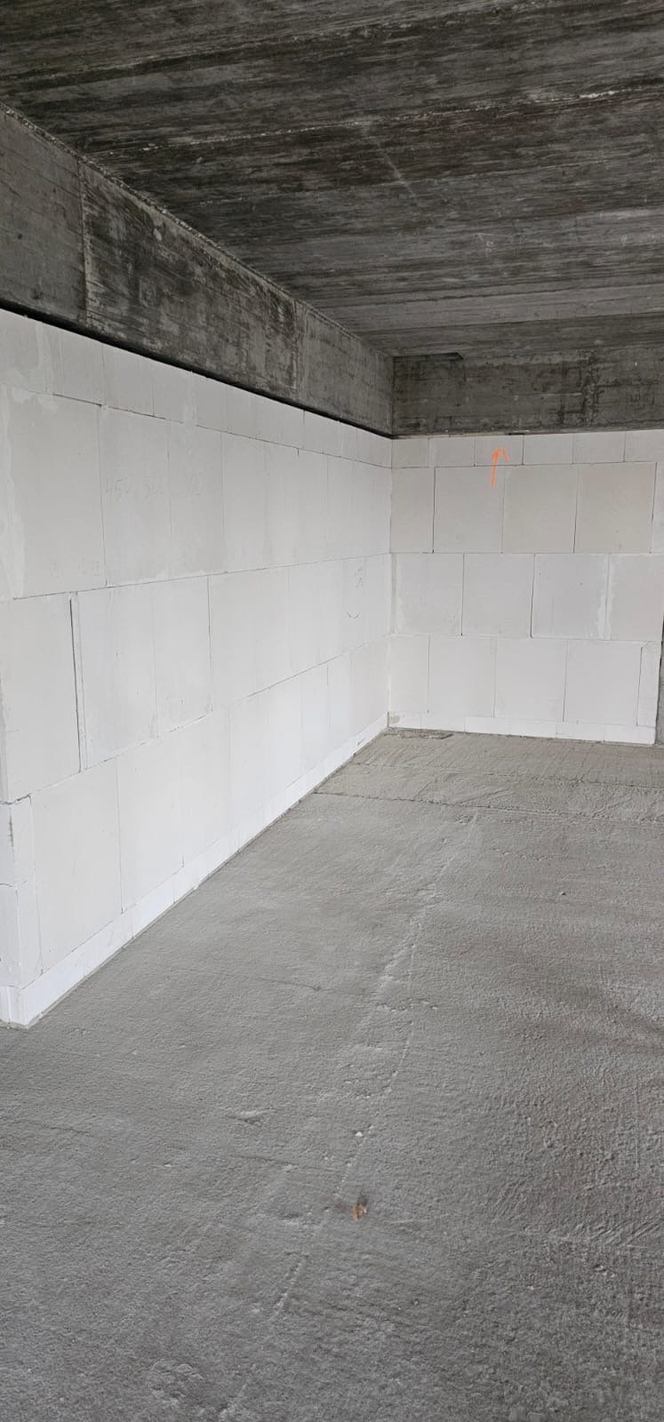 Wnętrze surowego domu w trakcie budowy z białymi ścianami z betonu komórkowego i surowym betonowym stropem, widoczny fragment pomarańczowej linii na ścianie.