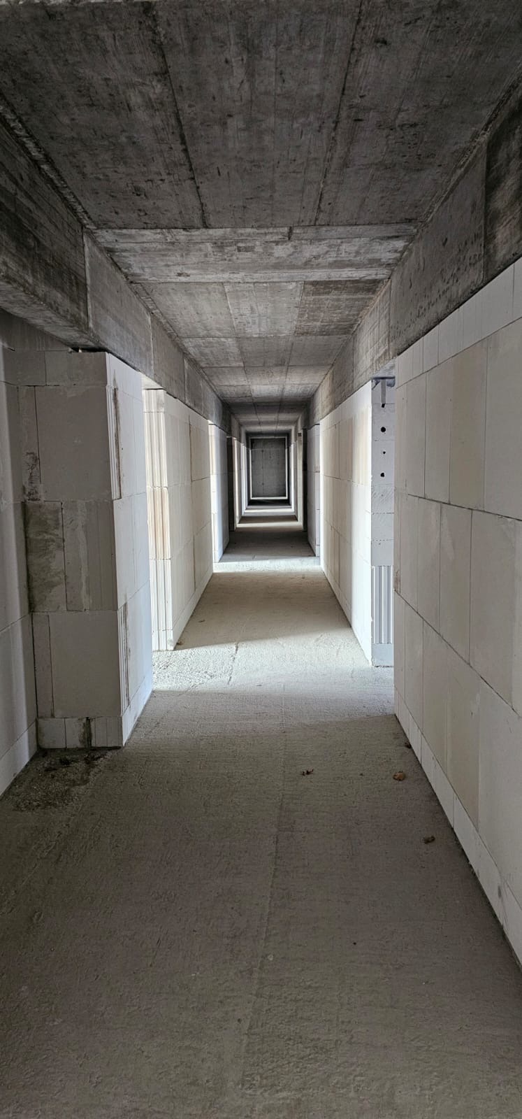Wnętrze surowego, długiego korytarza w budynku w trakcie budowy. Ściany z białych bloczków, surowy betonowy strop. Perspektywa zbieżna w głąb, światło dzienne wpadające z otworów po bokach.