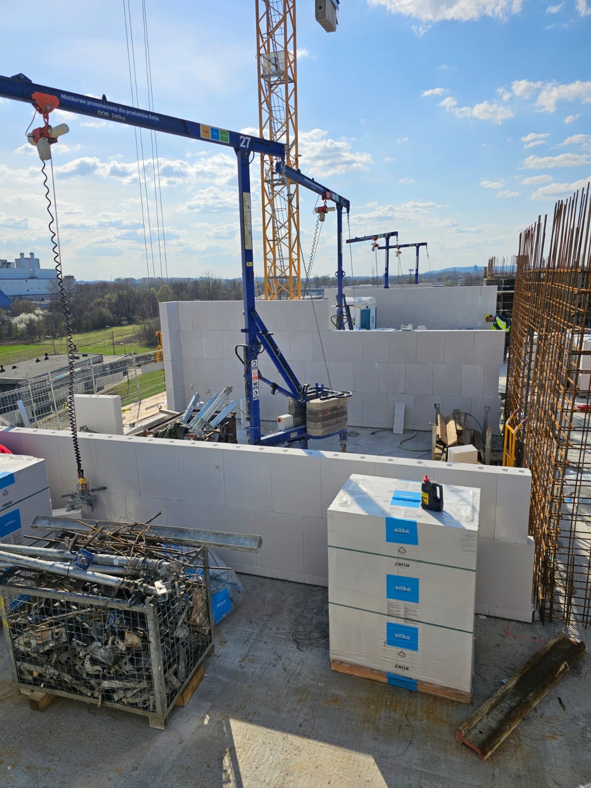 Budowa z bloczków Silka Xella: wznoszenie ścian z użyciem miniżurawia. Widoczne palety bloczków, stalowe zbrojenia i fragmenty konstrukcji budynku w tle.