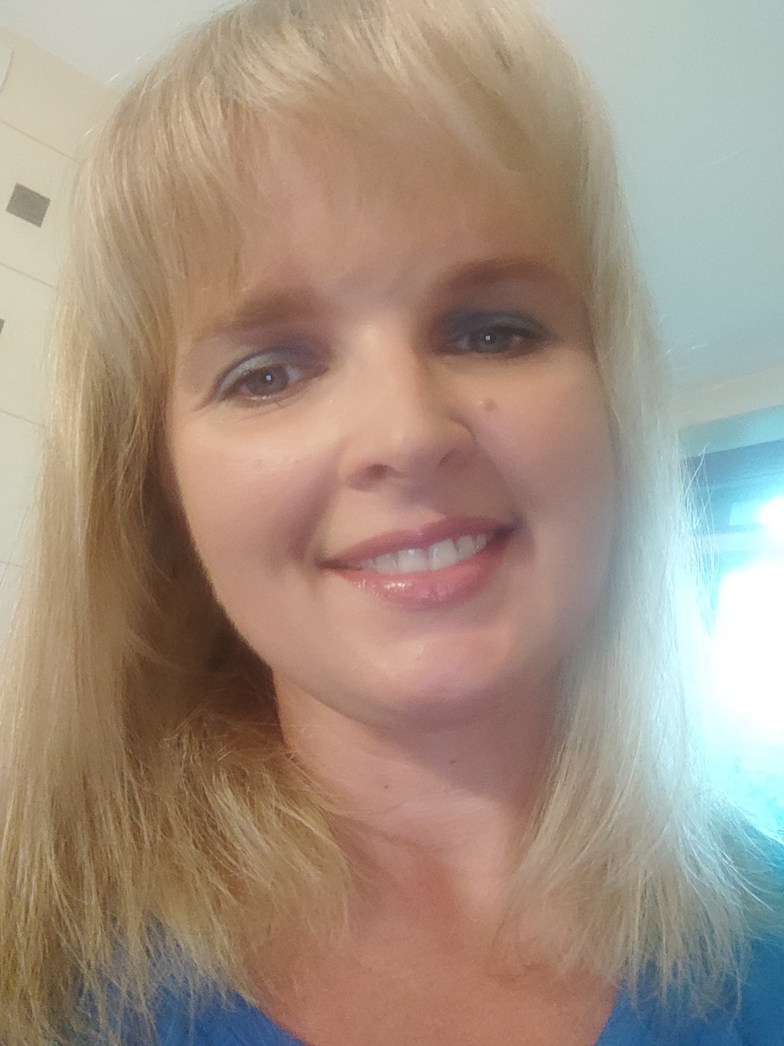 Portret uśmiechniętej blondynki z lekkim makijażem oczu, wykonany w jasnym wnętrzu.