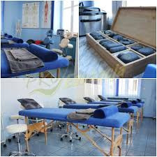 Wnętrze gabinetu z kilkoma niebieskimi leżankami do masażu, drewnianymi ramami i szarymi ręcznikami. W tle widoczne plakaty anatomiczne, sprzęt medyczny i drewniana skrzynia z kamieniami do masażu.