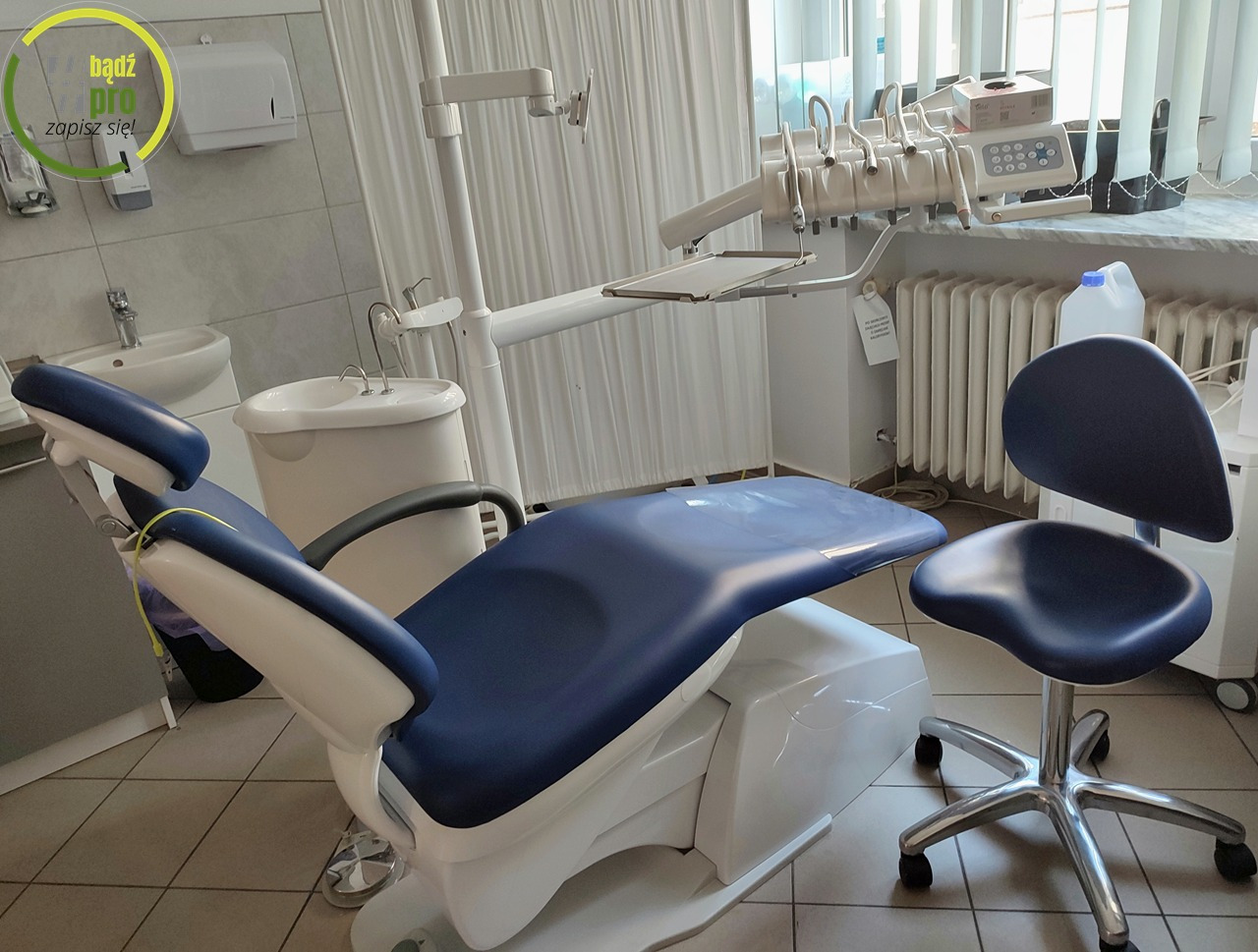 Wnętrze gabinetu stomatologicznego: fotel dentystyczny w kolorze granatowo-białym, obok krzesło na kółkach, w tle unit dentystyczny i umywalka.