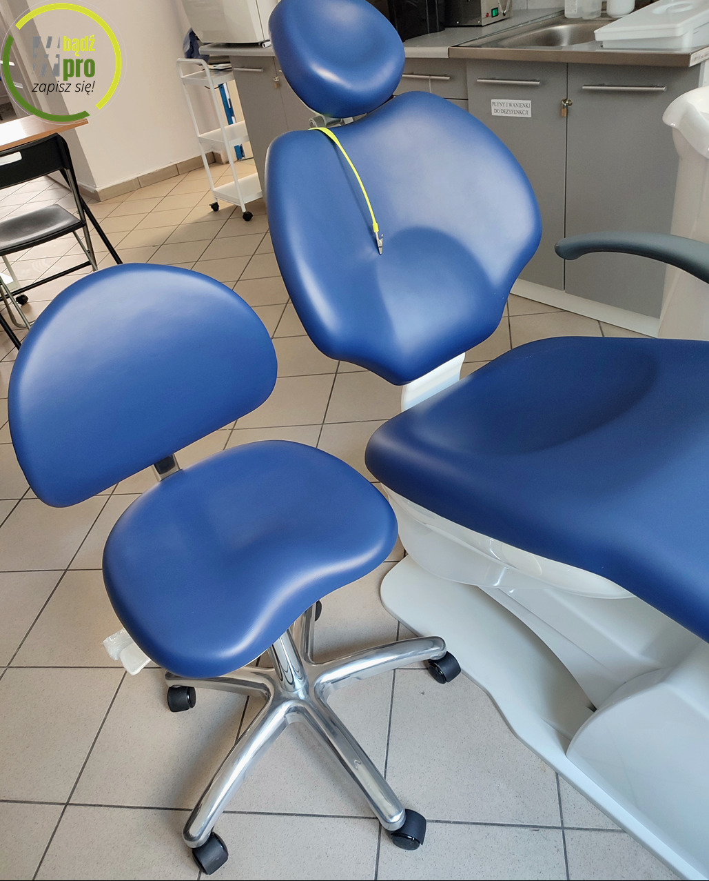 Niebieski fotel dentystyczny z regulowanym zagłówkiem i krzesłem dla lekarza w gabinecie w Katowicach, widoczne szafki i umywalka w tle.