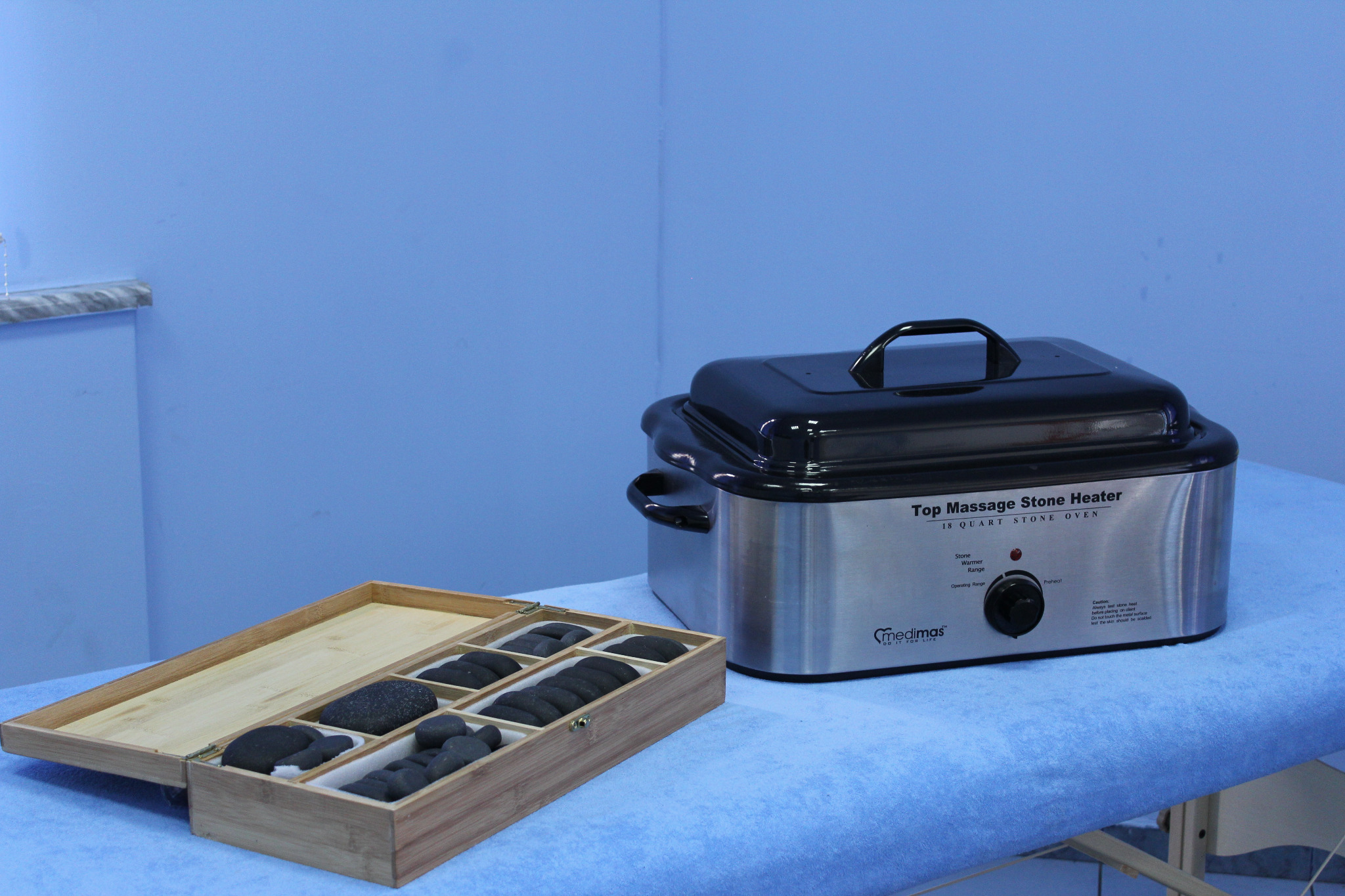 Podgrzewacz do kamieni do masażu z czarną pokrywą i metalową obudową, obok drewniane pudełko z przegródkami zawierającymi czarne kamienie do masażu na niebieskim tle.