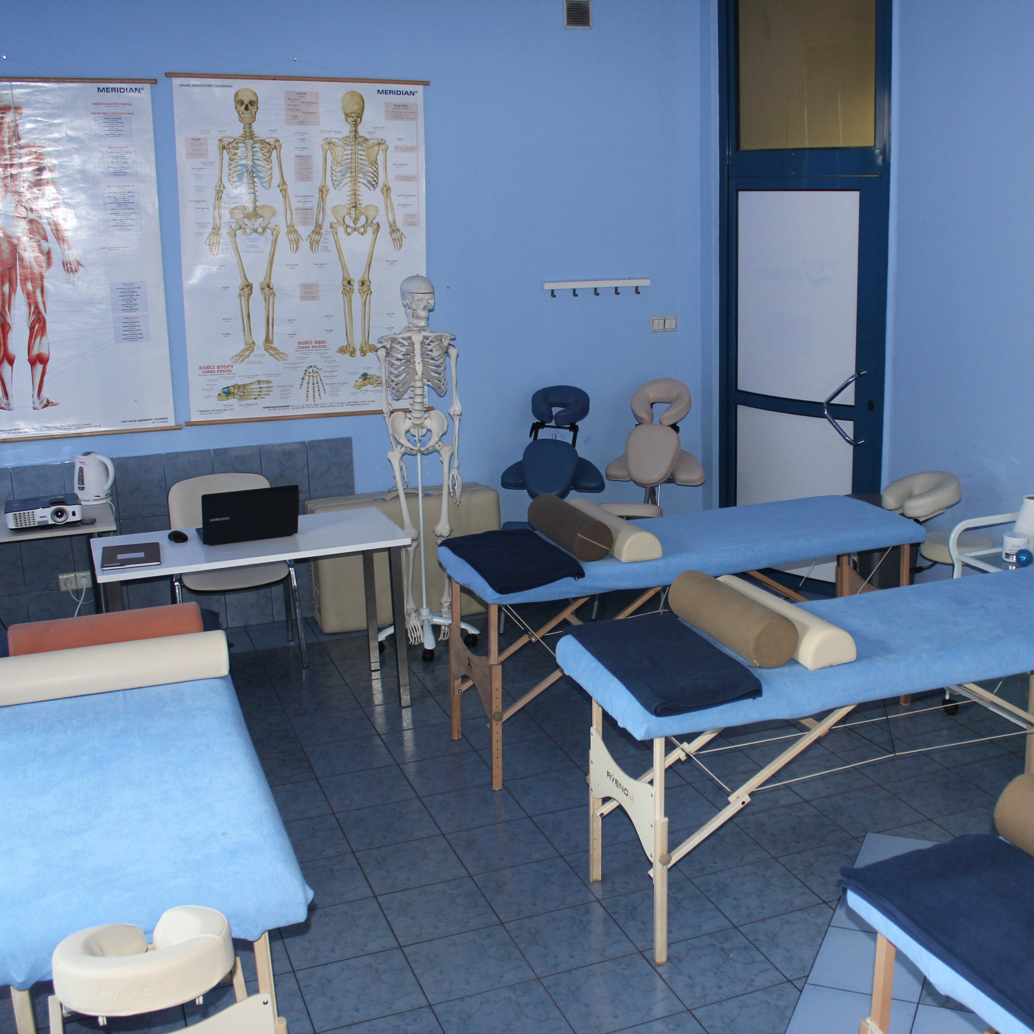 Sala szkoleniowa z niebieskimi ścianami, wyposażona w trzy leżanki do masażu z podgłówkami i wałkami, biurko z laptopem i projektorem, oraz plansze anatomiczne i model szkieletu.