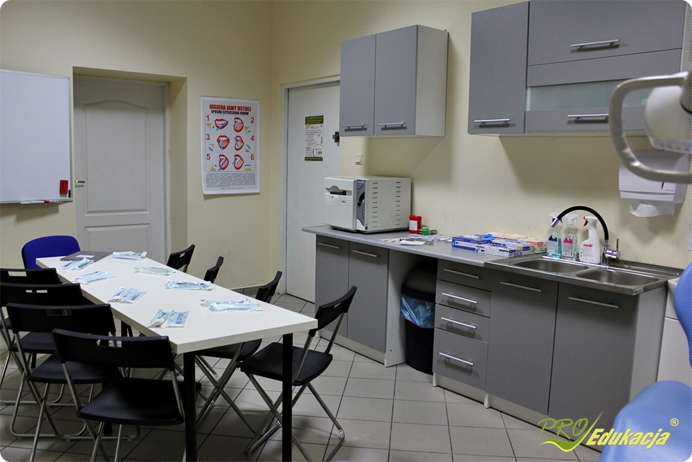 Sala szkoleniowa z plakatem higieny jamy ustnej, stołem z przygotowanymi pakietami i stanowiskiem do mycia rąk.