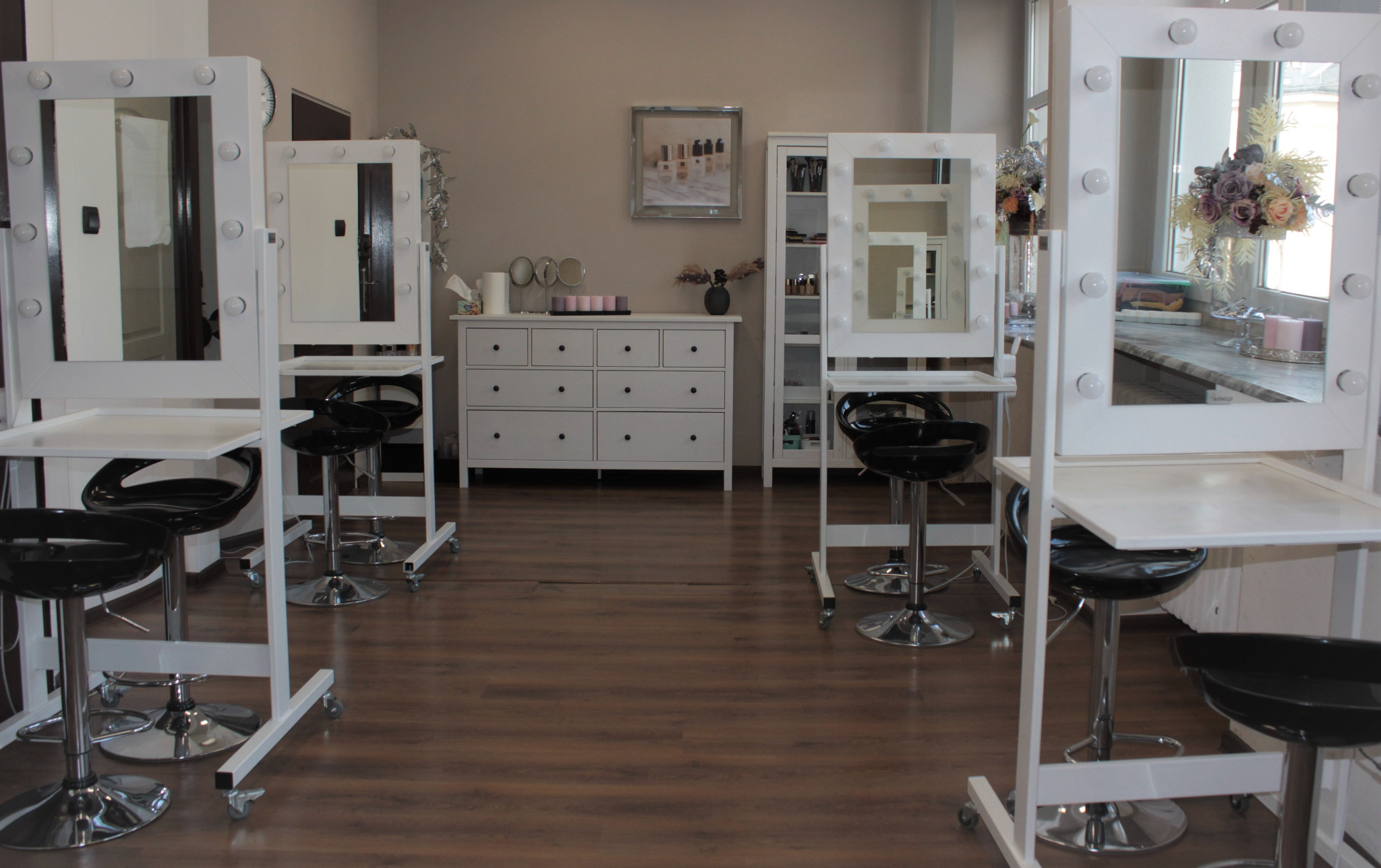 Wnętrze studia wizażu z czterema stanowiskami: białe toaletki z lustrami podświetlanymi żarówkami, czarne hokery na chromowanych nogach, drewniana podłoga, w tle komoda i regał z kosmetykami...