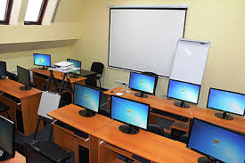 Sala komputerowa z rzędami biurek, na każdym biurku monitor z włączonym ekranem startowym Windows, rzutnik na statywie, tablica suchościeralna z markerami, widok z perspektywy ucznia.