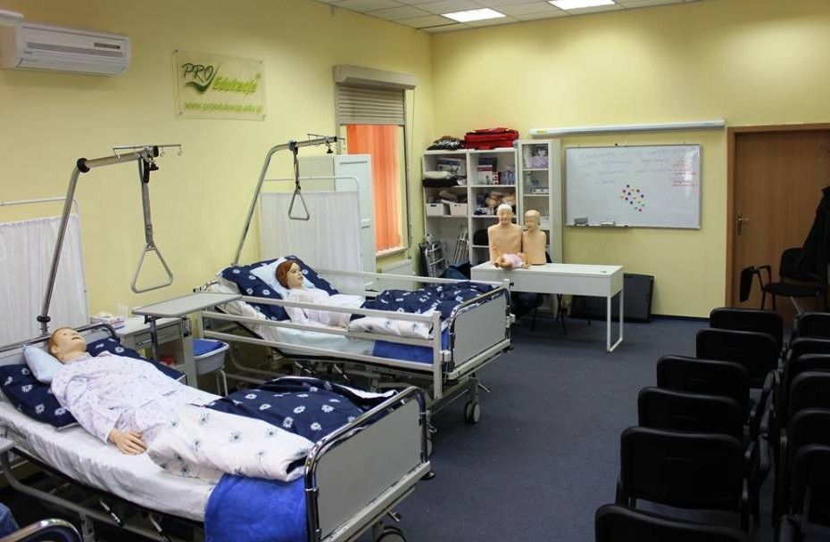 Sala szkoleniowa z dwoma łóżkami szpitalnymi z manekinami, tablicą i krzesłami, przygotowana do kursu pierwszej pomocy.