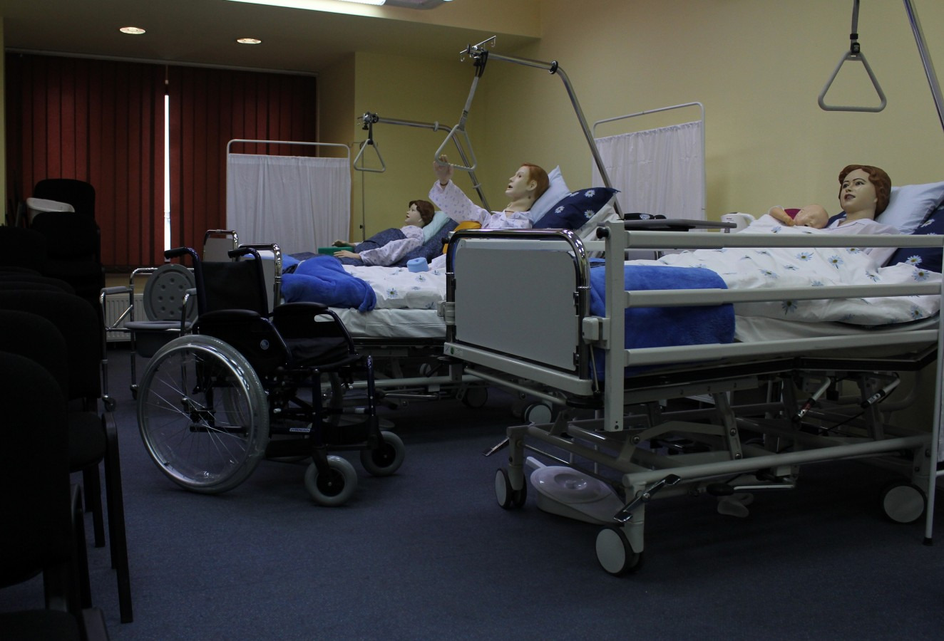 Sala szkoleniowa z łóżkami szpitalnymi, manekinami pacjentów, podnośnikami i wózkiem inwalidzkim w tle, imitująca warunki szpitalne.