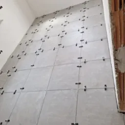 Ściana w trakcie układania szarych płytek, widoczne krzyżyki dystansowe i fragment ściany z cegły.