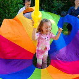 Dziewczynka w różowym stroju bawi się z uśmiechem na twarzy, trzymając żółty balon w kształcie miecza, podczas gdy dwóch chłopców trzyma kolorową chustę animacyjną z otworami w ogrodzie.