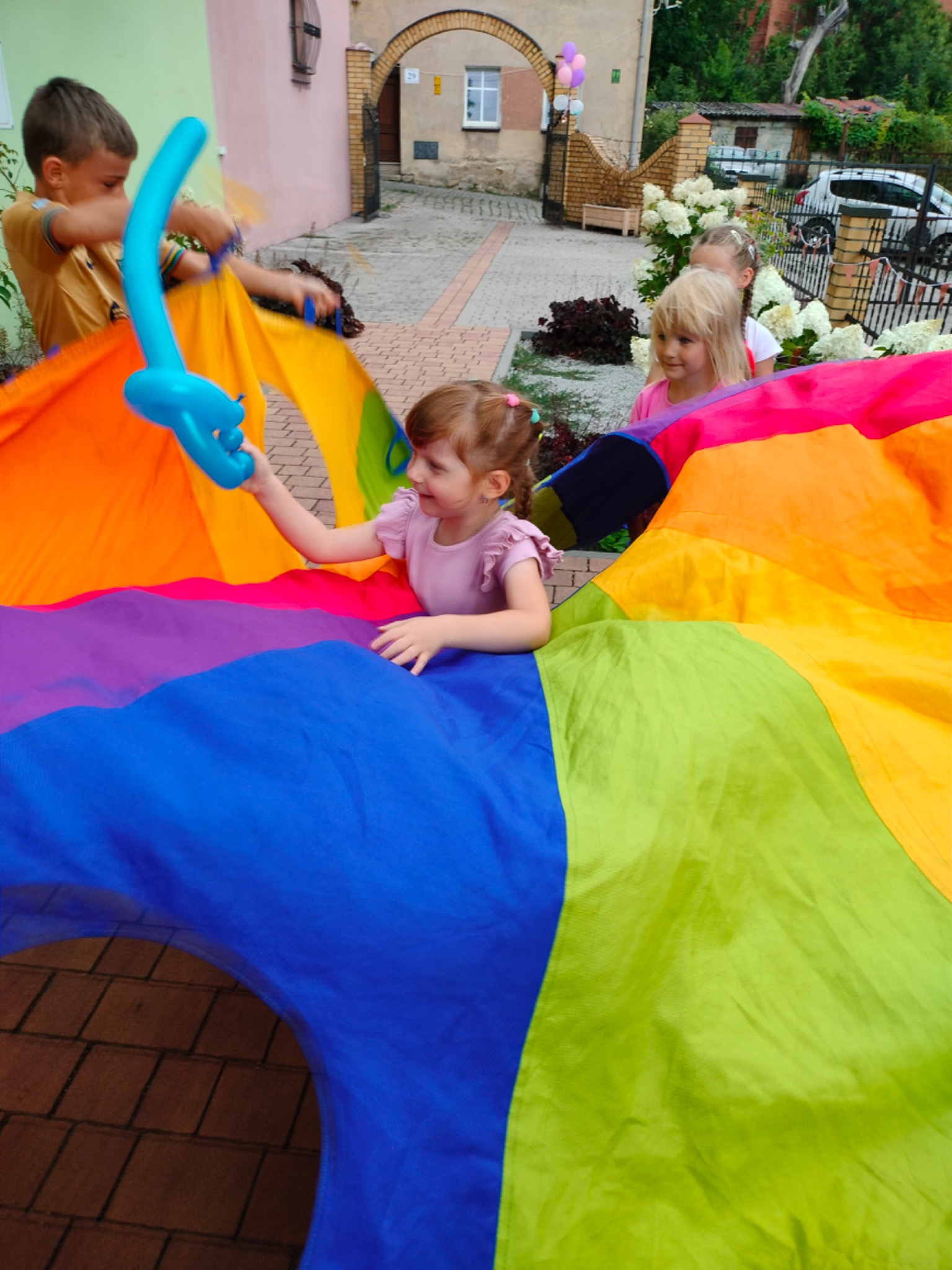 Dzieci bawią się kolorową chustą animacyjną na zewnątrz, jedno z nich trzyma niebieskiego balonowego pieska.