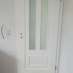 Białe drzwi wewnętrzne z dwoma pionowymi szybami z matowego szkła, widoczne zawiasy i zamek, na ścianie gniazdko elektryczne z wystającym przewodem, parkiet na podłodze.