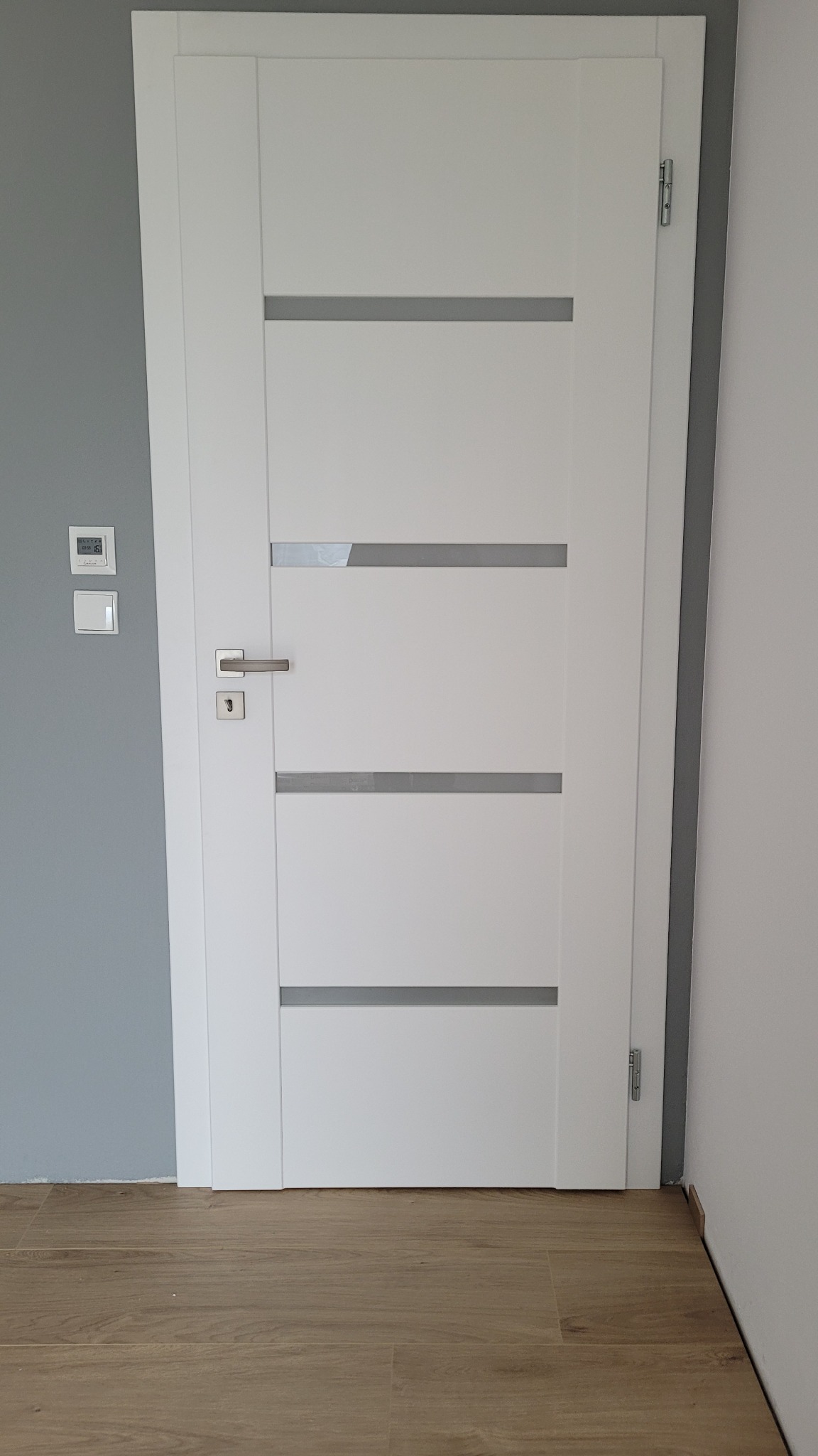 Białe drzwi wewnętrzne z czterema poziomymi, szarymi wstawkami, srebrna klamka, widoczny fragment szarej ściany z termostatem, podłoga imitująca drewno.