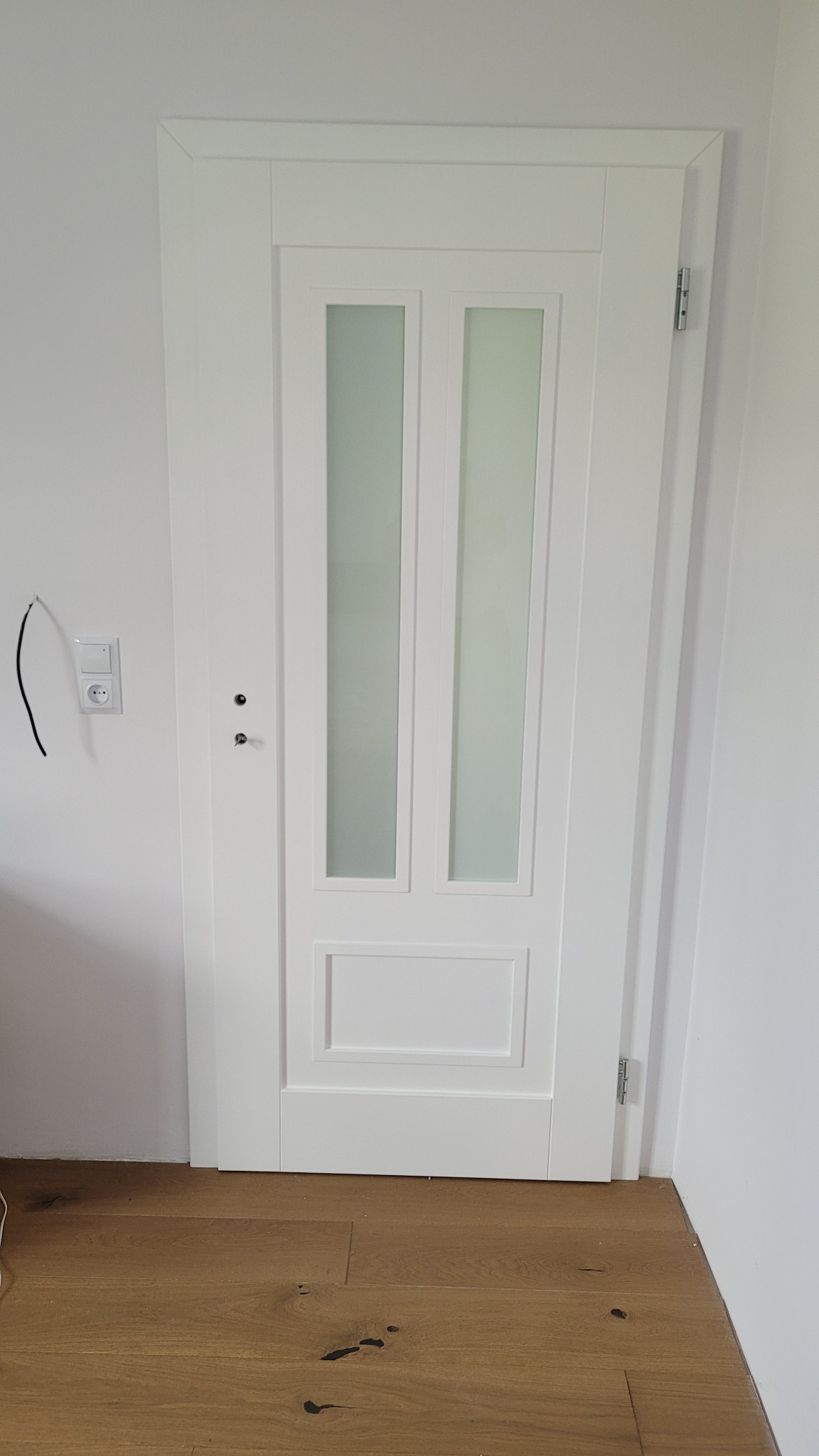 Białe drzwi wewnętrzne z dwoma pionowymi szybami z matowego szkła, widoczne zawiasy i zamek, na ścianie gniazdko elektryczne z wystającym przewodem, parkiet na podłodze.