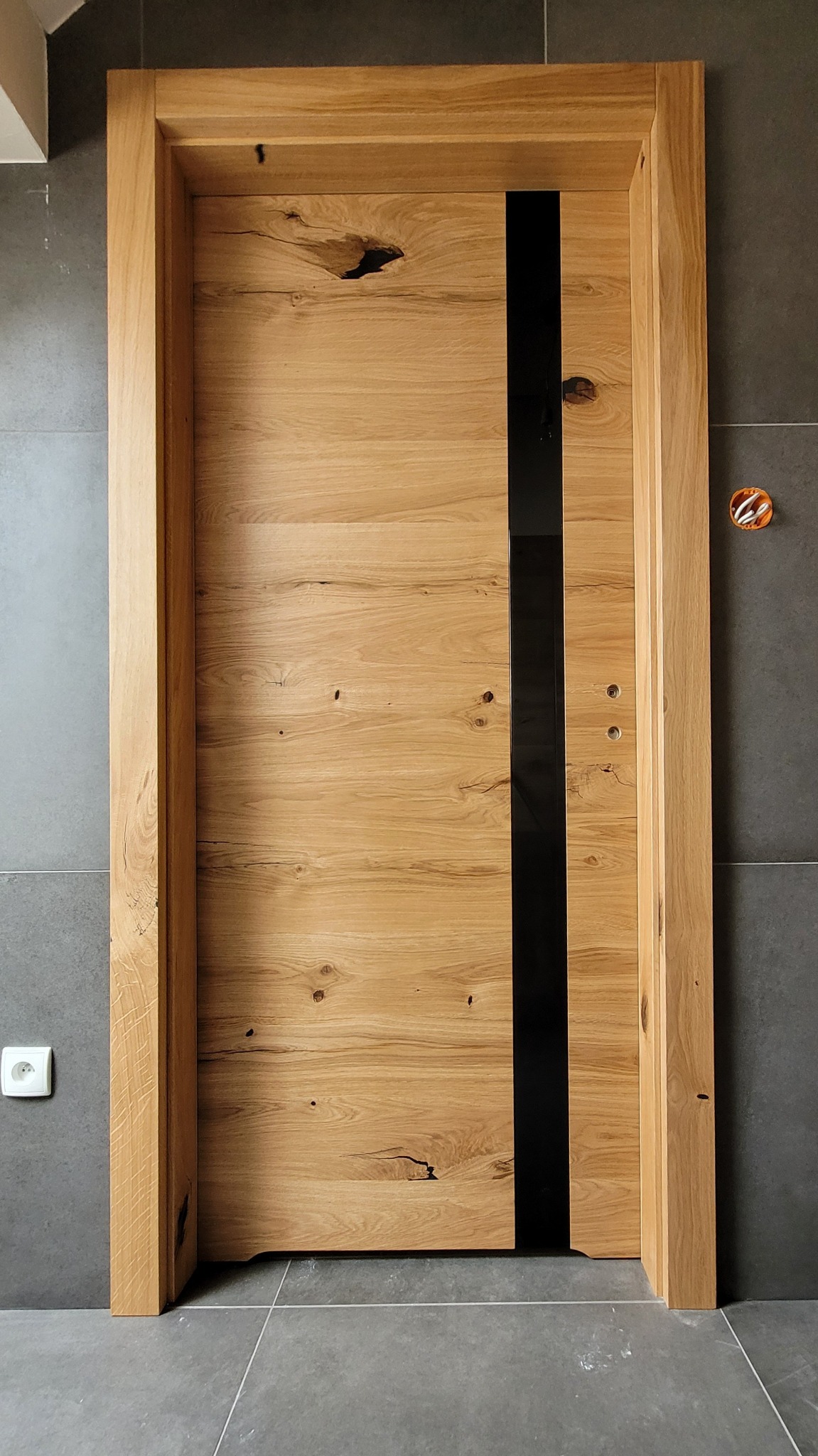 Dębowa ościeżnica i skrzydło drzwiowe z pionowym, czarnym pasem na tle szarej, matowej ściany i podłogi z płytek, widoczna instalacja elektryczna w ścianie.