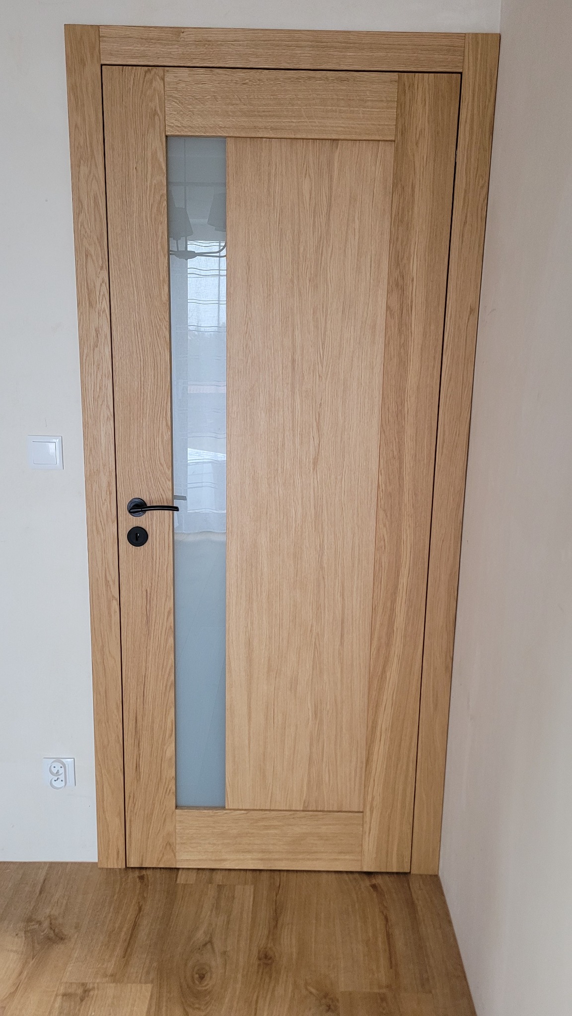 Dębowa ościeżnica z czarną klamką i pionowym przeszkleniem wstawiona w pomalowaną na biało ścianę, widoczny fragment parkietu.